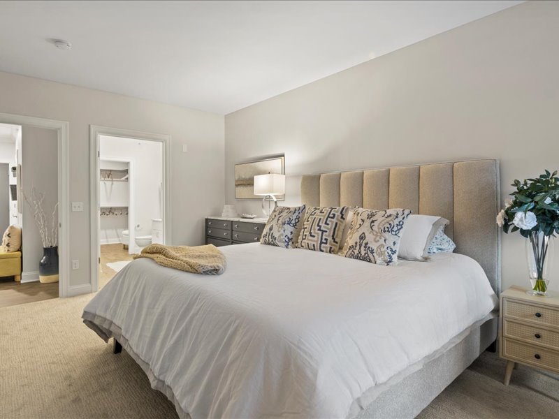 The bedroom has a king-size bed with a white comforter and pillows. There is a nightstand next to the bed with a bocque of flowers. There is also a dresser against the wall opposite the bed. The bedroom has a neutral color scheme with white walls and light wood furniture.